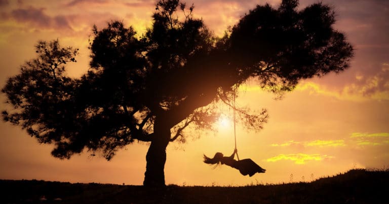 A silhouette of a girl releasing things to let go of while swinging on a tree at sunset.