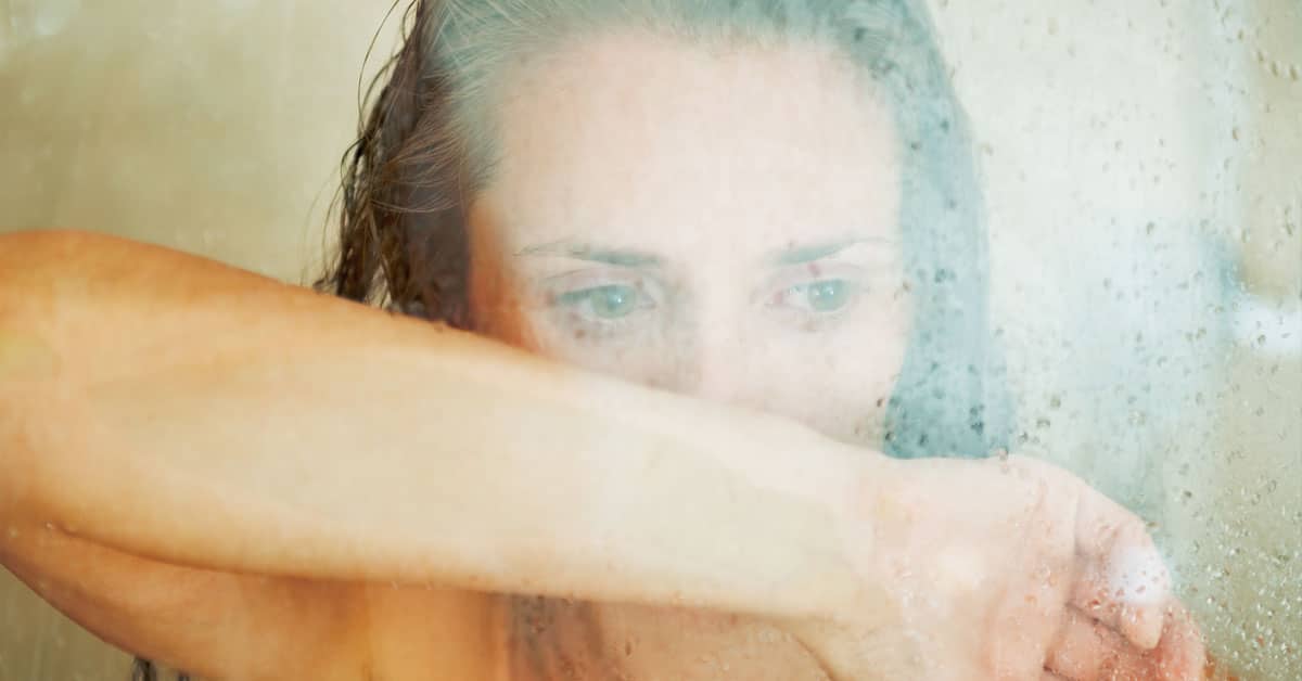 A woman appears discontent as she takes a shower, her hand aimlessly submerged in the water.