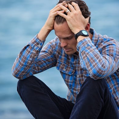 An overstressed man sitting on the beach with his hands on his head.