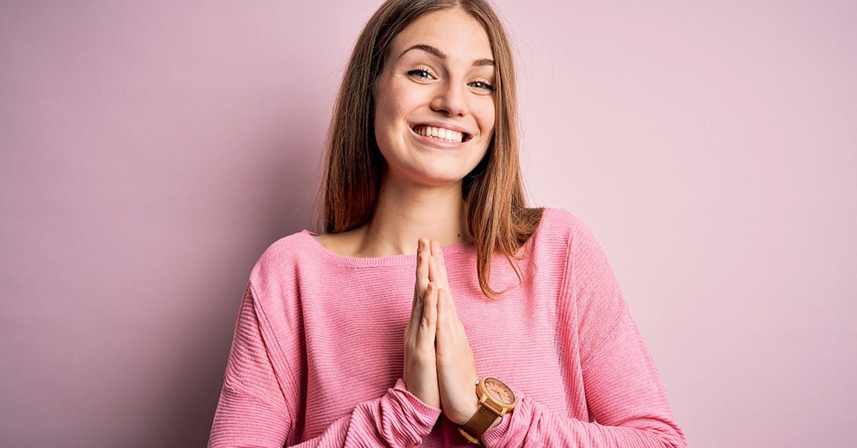 A young woman with her hands folded in prayer on a pink background, embodying self-kindness.