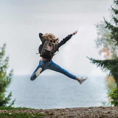 A joyous woman leaping with a backpack by the ocean.