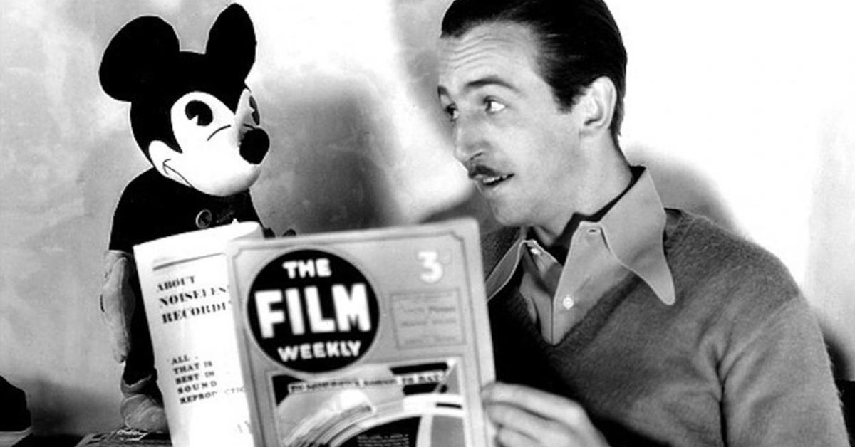 A man reading a book with a mickey mouse stuffed animal.