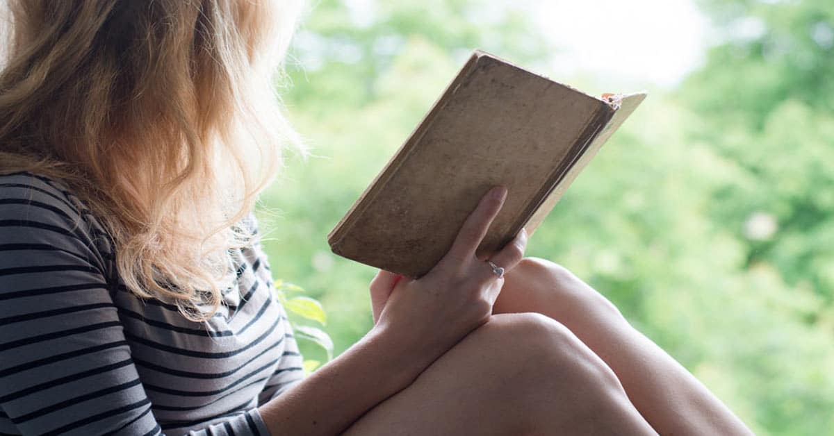 A woman gaining lessons on life while sitting on a window sill reading a book.