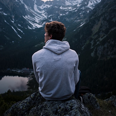 Young man sitting on a rock cliff looking out into the mountain landscape