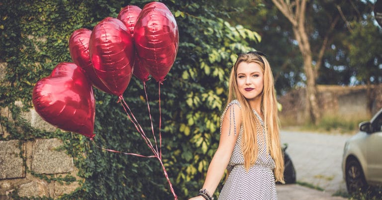A girl displaying festive red heart balloons while standing beside a car.